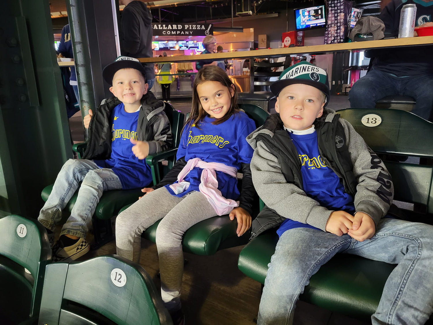 Three children in sports-themed shirts sitting in a stadium seat, with a sports arena in the background.